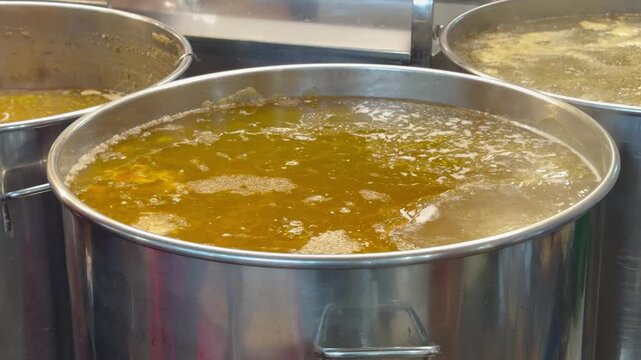 Large stainless steel pots filled with hot broth or soup cook indoors in a professional kitchen. Catering service, commercial cooking, restaurant food preparation.