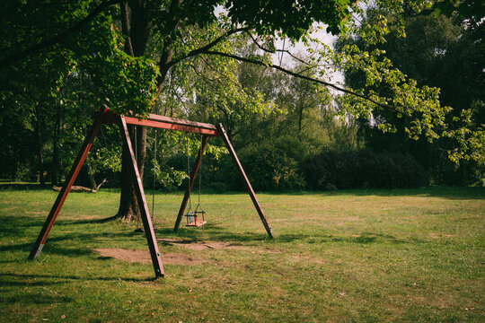 Empty wooden swing set in a grassy park under dappled tree shade evoking childhood nostalgia and vintage outdoor recreation themes for editorial design