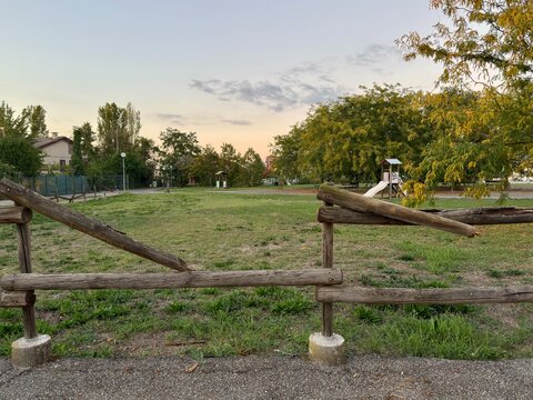 Broken wooden fence surrounding empty public park playground at sunset. Damaged timber railing creates safety hazard requiring municipal maintenance and repair.
