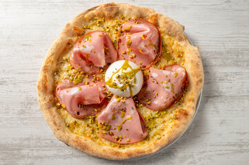 Overhead view of a gourmet Italian pizza topped with mortadella, creamy mozzarella and chopped pistachios on a light wooden surface. © Lorenzo Dottorini