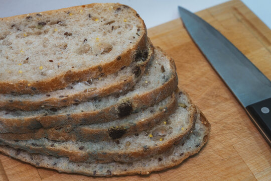 rotten bread pieces exhibiting fungal mold growth stay arranged on wooden cutting board near chef knife. decayed multigrain loaf pieces illustrate inedible stale bakery contamination closeup.