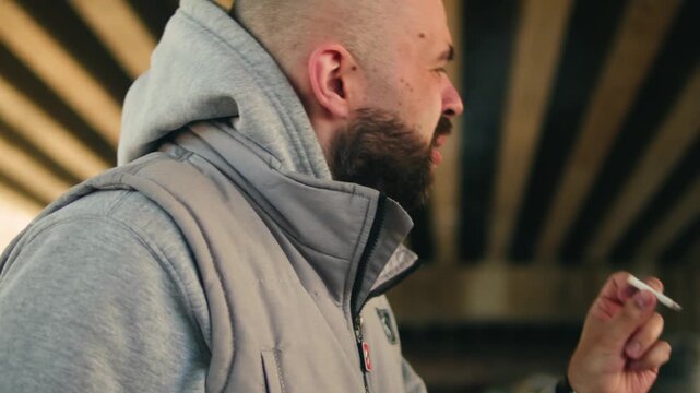 Man smoking vape and cigarette outdoors, bearded male inhaling electronic cigarette smoke under bridge, nicotine addiction and bad habit concept close up