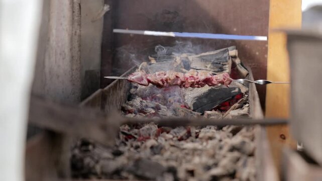 Meat skewers roast over charcoal as smoke rises from glowing embers. Grilled meat cooks above fire, with small flames flickering beneath metal spit in open hearth, while coals glow below.