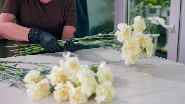 Florist sorting white carnations in flower shop, woman hands in black gloves preparing cream carnation bouquet, floristry small business close up