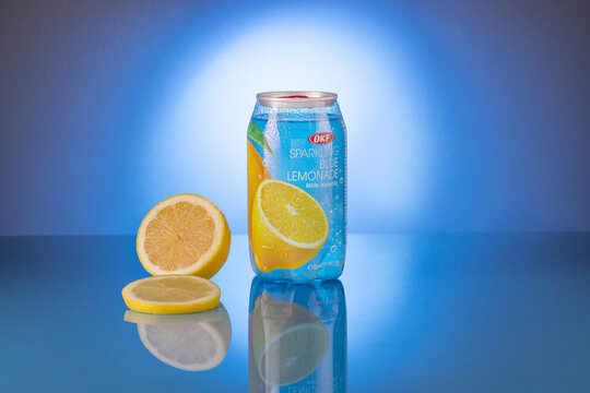 OKF Sparkling Blue Lemonade in a transparent container with condensation, beside a lemon half and slice on a reflective blue surface against a blue studio background, with copy space.