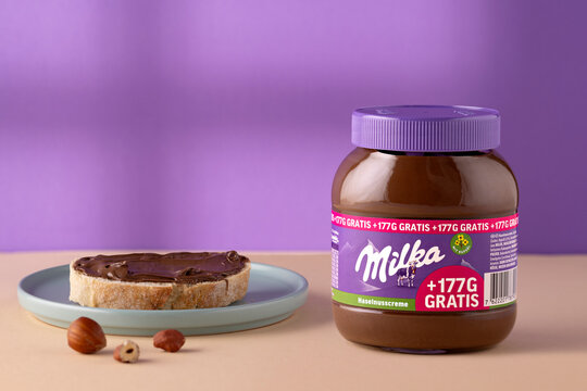 Milka Haselnusscreme hazelnut cream jar beside bread topped with chocolate hazelnut spread on a ceramic plate, with hazelnuts on a cream-colored surface against a lavender background, with copy space