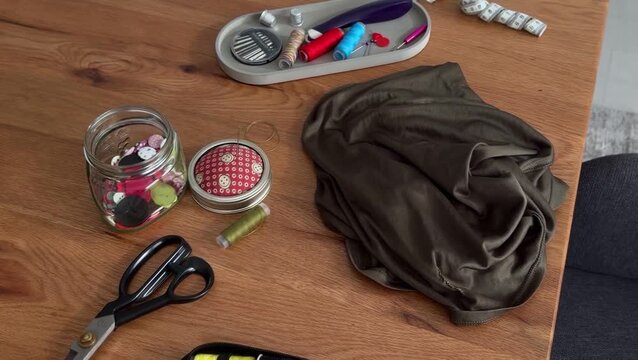 Sewing supplies and folded fabric arranged on a wooden table with scissors, colorful thread spools, measuring tape, buttons, and a small notions tray