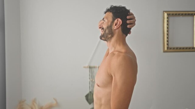 Shirtless man standing with hand behind head showing bare chest in a bedroom studio with macrame wall hanging and empty picture frame; serenity.