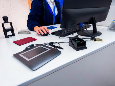 A Ukrainian civil servant or office worker processes a biometric passport registration at a desk equipped with an electronic signature pad, fingerprint scanner, and stamps (close-up). Ukrainian passpo