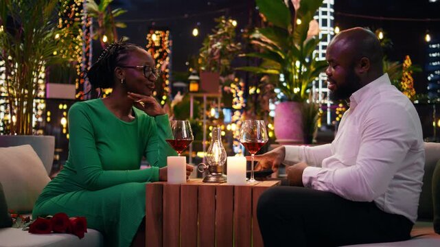 Happy man and woman use a phone to take pictures during a romantic rooftop restaurant date under glowing lights. Joyful girlfriend and boyfriend take selfies together with a mobile phone.