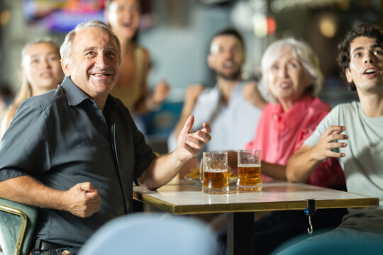 Elderly man is sitting at a table in a bar against the background of a group of friends, they are actively watching the game and football