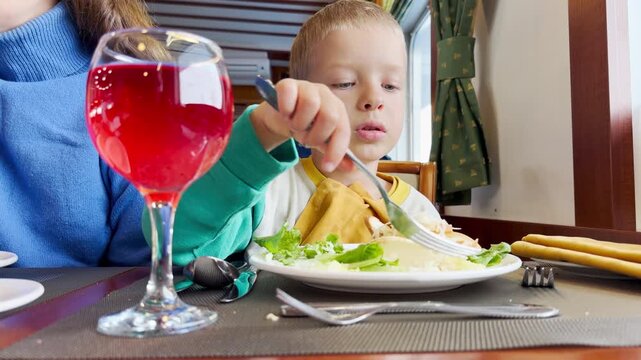 A happy boy reaches with a fork to eat his fresh salad. He dines with joy inside a warm cruise ship restaurant.
