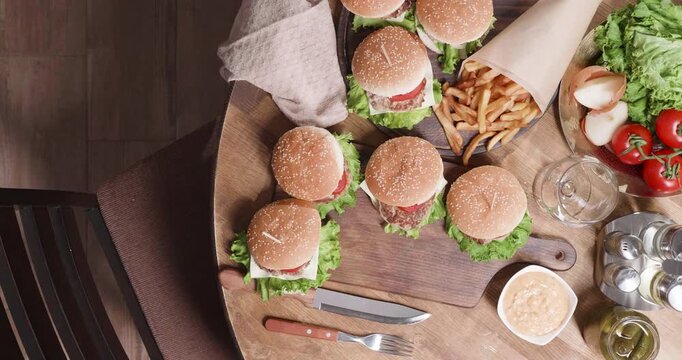 Burgers, hands and serving from an overhead view as a person arranges a cheeseburger among fries, tomatoes, lettuce, pickles and sauce on a wooden table for casual dining and food preparation.