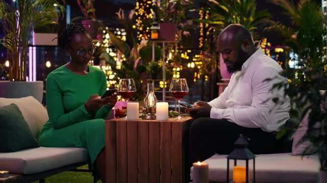 Bored couple uses a mobile phone and avoids conversation during date night, while the man and woman neglect each other at a rooftop restaurant dinner and browse smartphones instead of talking.