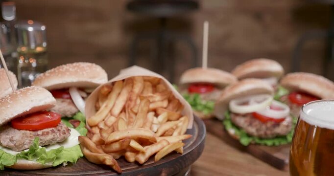 French fries, burgers and rotation on a dark wooden serving board, with golden fries spilling from a paper cone beside sesame bun cheeseburgers and beer in a rustic pub-style food presentation.