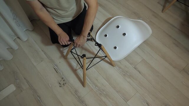 Overhead view of person assembling modern wooden leg chair, using Allen key to tighten metal support, align wooden legs, while loose screws and washers rest