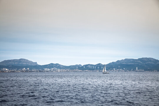 A sailing boat crosses the Mediterranean Sea off Marseille, France, with white sails raised against a panorama of waterfront buildings and rugged hills stretching along the city's distant coastline.