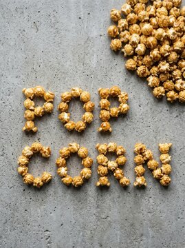 High angle view of sweet golden caramel popcorn kernels arranged to spell the word POP CORN on a textured gray concrete background, a popular delicious snack for movie nights and entertainment.