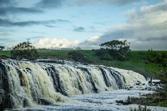 Wide waterfall cascading over dark rock formations, under dramatic, cloud-filled sky, surrounded by green countryside, with people fishing at bottom of fall, Hopkins Falls, Cudgee, Victoria, Australia