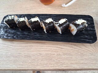 Japanese sushi rolls wrapped in nori seaweed and topped with mayonnaise, served on a black rectangular plate on a wooden table. © putri