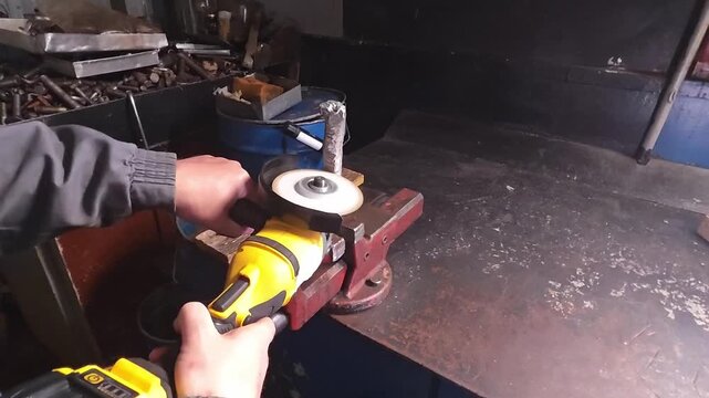 Angle grinder is aligned on secured metal workpiece at bench vise. Worker steadies yellow tool on work surface, concentrating on precise placement inside busy workshop with scattered scrap.