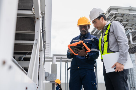 Diverse professional industrial maintenance team inspecting rooftop HVAC cooling systems using a rugged digital tablet during a routine commercial facility infrastructure audit.