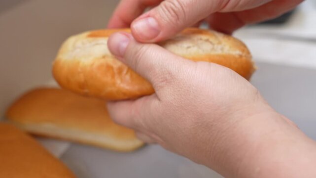 Hands cut a soft golden hot dog roll lengthwise with a knife, then pull it open to prepare a sandwich in a kitchen while other buns rest in a box nearby.