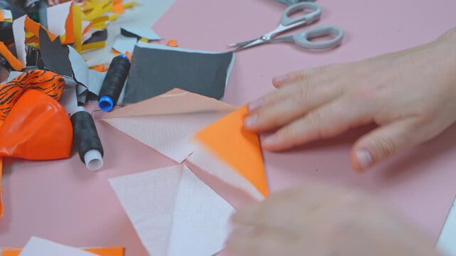 Woman hands move triangular white and orange fabric cutouts across a pink worktable, beside black thread spools, scissors and scraps, a handmade sewing and patchwork project at home.
