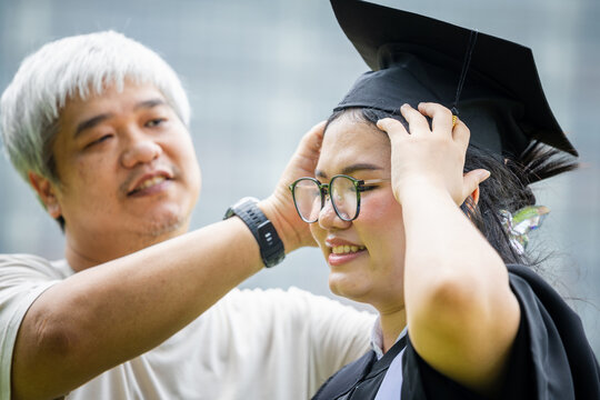Asian father helping daughter adjust graduation cap on university commencement day, Loving parent assisting graduate daughter with academic cap during graduation ceremony