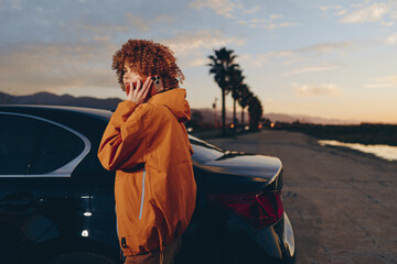 Naklejka premium Woman lifestyle outdoors near car at sunset, hooded jacket in warm orange, standing on road with distant mountains, one hand touching hair, calm travel mood, soft cinematic light today
