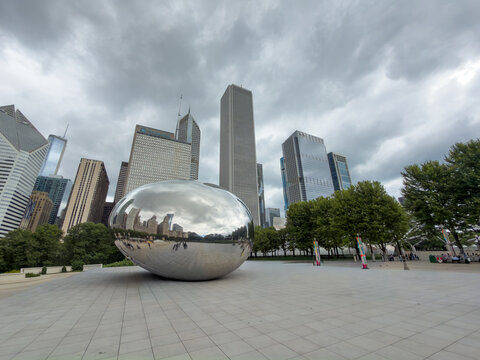CHICAGO, IL, JUN 18, 2026: Wide-angle view of Cloud Gate on its stone plaza in Millennium Park, with Chicago skyscrapers, green trees and dramatic storm clouds overhead