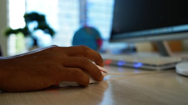 Hand using a computer mouse at a modern workspace, Close-up of a hand using a computer mouse on a wooden desk, with a blurred monitor and home office background, suggesting digital work, browsing.