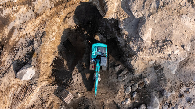Aerial view of an excavator working in a deep dirt pit with rocky terrain and shadows cast on the ground in Indonesia