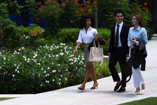 Three young colleagues walking past a hedge of white flowers on a bright morning in a modern mixed use development, navy suit with white and light blue shirts, easy everyday workday feeling