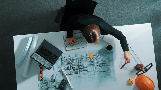 Aerial view of architect drawing while working at laptop. Top down view of caucasian engineer designing blueprint and drafting house model at table with equipment and safety helmet. Alimentation.