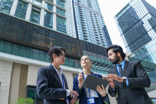 Three young business professionals wearing conference badges review data on a tablet in front of a glass tower beside the polished facade of a new mixed use city development