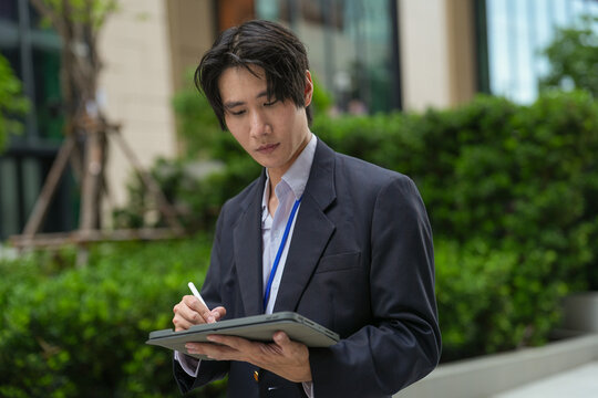 A focused young Asian executive considers a decision while cradling his tablet in front of luxury retail frontage in a central business district, modern towers rise steeply behind him