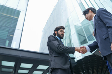 Two businessmen and a businesswoman welcome a partner with a firm outdoor handshake in the green outdoor courtyard of a downtown business complex, glass towers rise behind the handshake © ultramansk