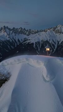 Vertical video: Gliding drone passing snow ridge at dusk, showing lantern post to capture peaks