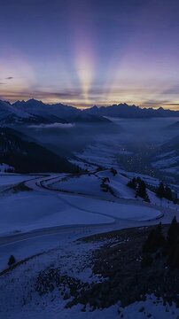 Vertical video: Moving camera revealing snow slopes and road toward valley at dawn, capturing rays