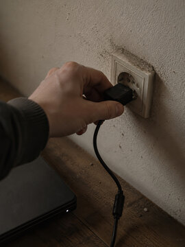 A man is connecting a black cable to a white electrical outlet on a textured wall in a dimly lit room.
