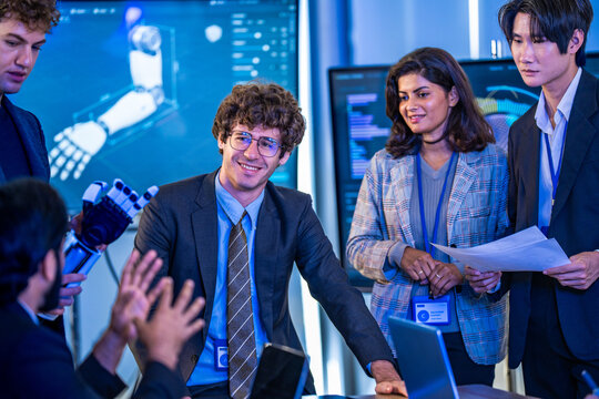Young engineer in suit smiles while bearded colleague claps holding a robotic hand, Indian colleague with reports behind, in a documentary-style moment, a digital transformation concept