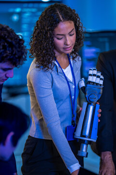 Curly-haired female engineer with lanyard holds a silver bionic robotic arm as colleagues lean in, in a real working moment, a scene of bionic prosthetics innovation