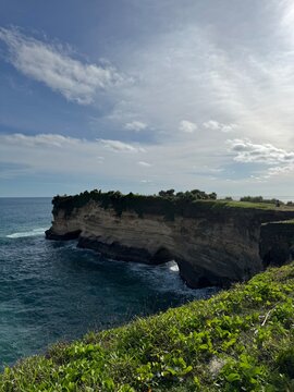 Dramatic limestone sea cliff topped with green grass and shrubs overlooks a natural rock arch where ocean waves crash below, with clear blue sea stretching to the horizon under a partly cloudy sky. Lu
