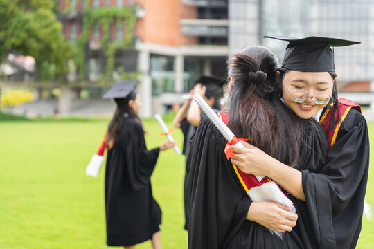Asian female graduate hugging classmate with diploma scroll on university campus, Graduates embracing warmly with diploma certificates on green campus field
