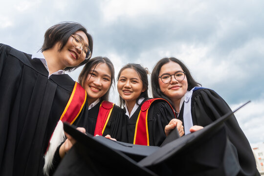 Group of cheerful university graduates in academic gowns smiling down at camera, Joyful asian students holding graduation caps and smiling together outdoors
