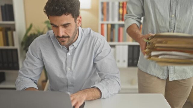 Man at desk handing a stack of folders and gesturing with his hand beside an open laptop while glancing aside in building; work stress.