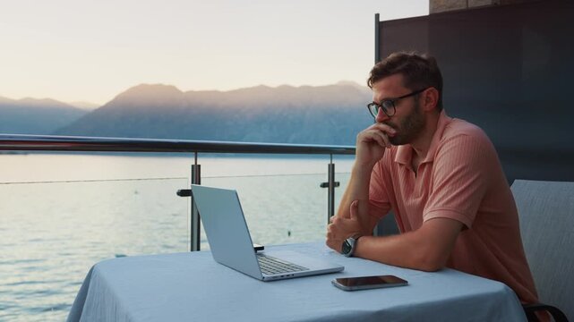 Focused remote worker using laptop on peaceful waterfront balcony at sunset, reflecting on tasks and enjoying serene outdoor workspace by calm scenic sea