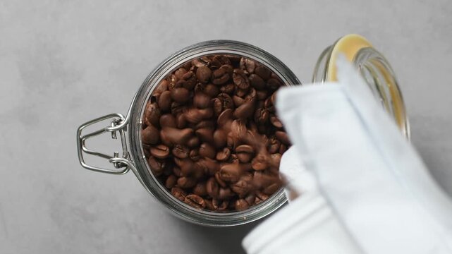 A person tops up a glass jar with roasted coffee beans, closes its clasp lid and lifts it away.