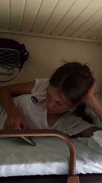 A teenage girl is holding a phone in her hand and sending a text message while sitting in her compartment during a train trip. The girl is playing online games while lying on her seat in the third cla